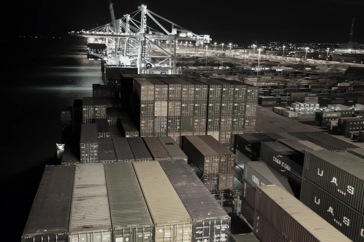 Rows of shipping containers stacked at a port at night, with gantry cranes lit behind them
